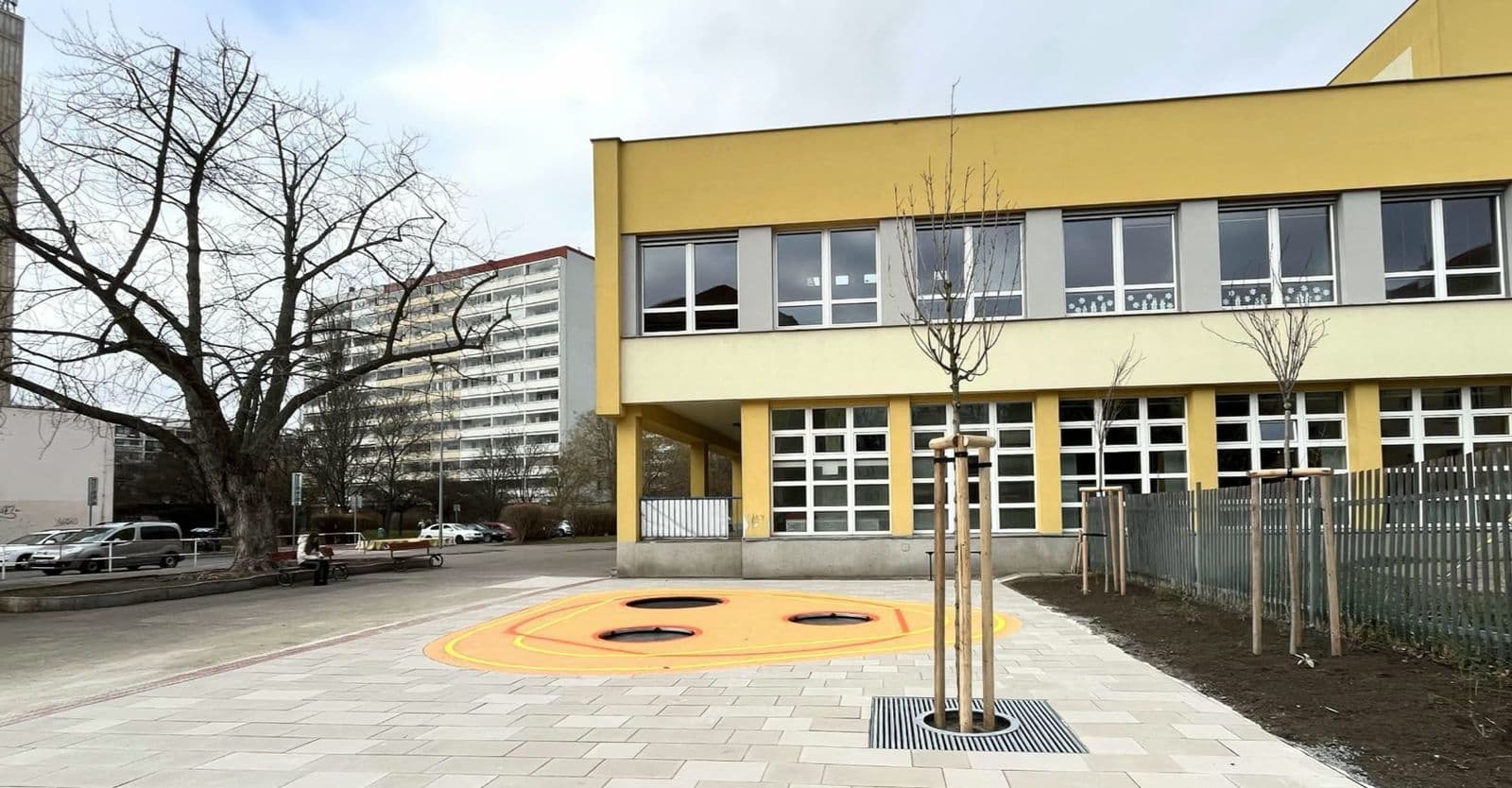 Školní dvůr u žluté budovy: nová dlažba, oranžová plocha s trampolínami, mladé stromy a čerstvě osázené záhony u plotu