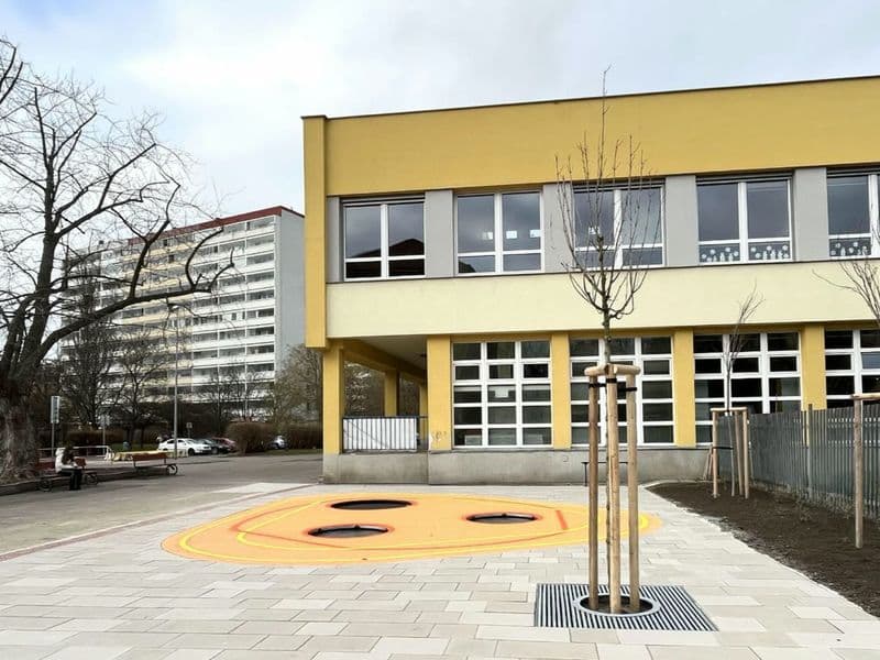 Školní dvůr u žluté budovy: nová dlažba, oranžová plocha s trampolínami, mladé stromy a čerstvě osázené záhony u plotu