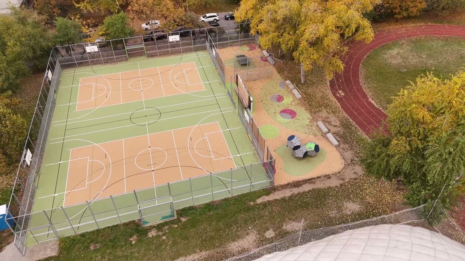 Nové multifunkční hřiště u školy z dronu: oplocené kurty, vedle hrací zóna, atletická dráha a podzimní stromy